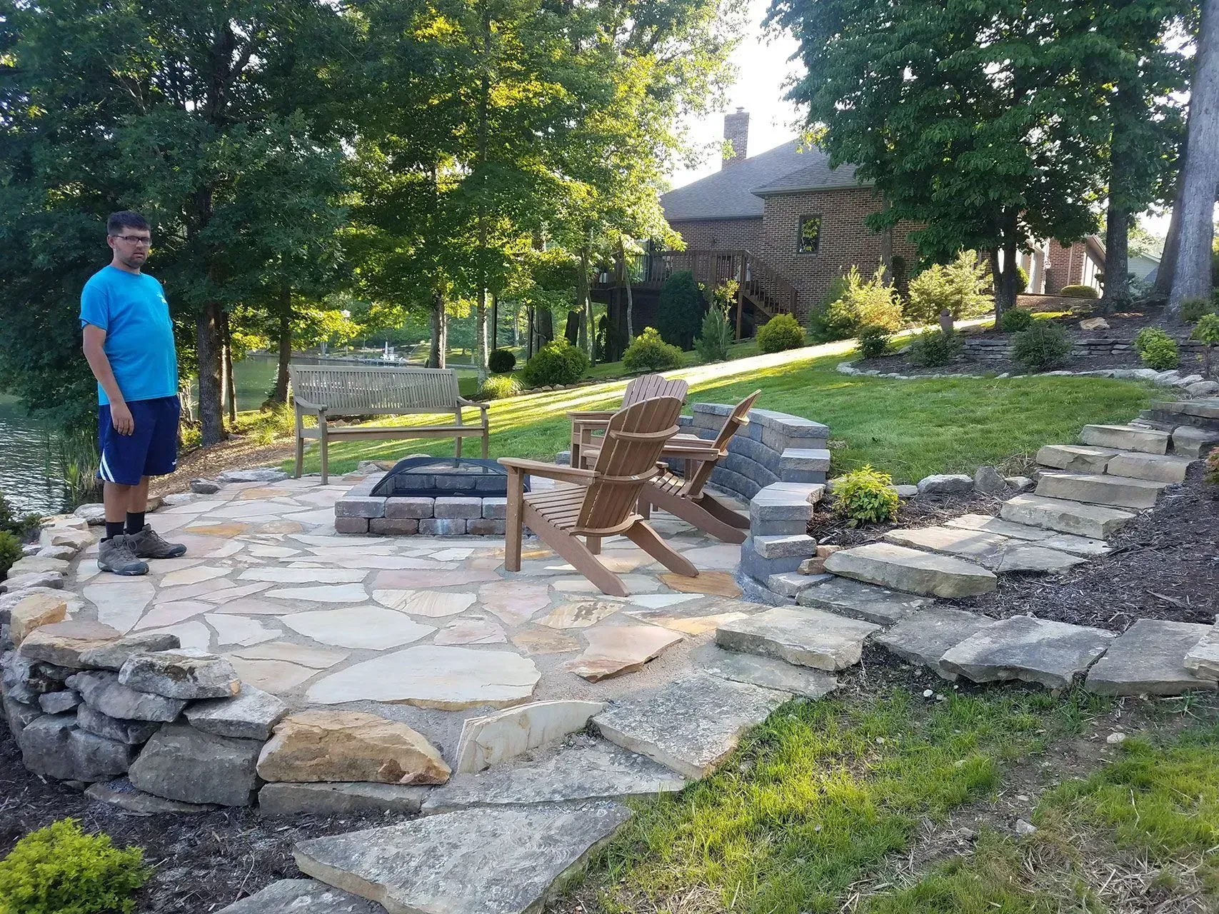 Man standing by a stone patio with Adirondack chairs and garden steps in a landscaped yard