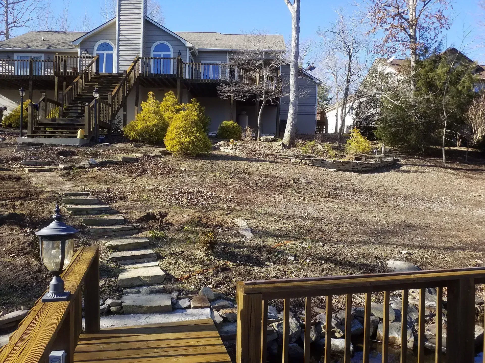 Backyard with stone steps, wooden deck, and gray house under a clear blue sky.