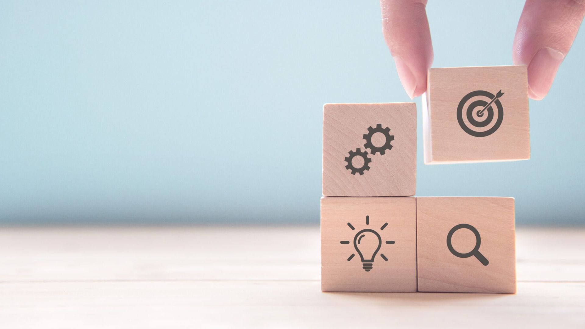 Hand placing a wooden block with a target icon on a stack of blocks with gears, lightbulb, and magnifier icons