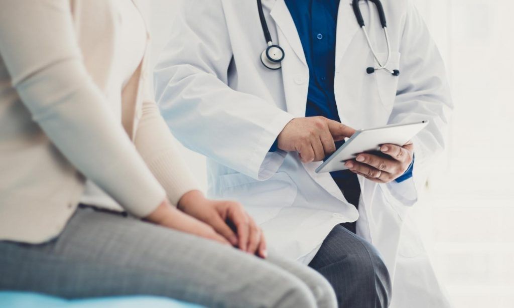 Doctor in white coat reviews tablet with seated patient.