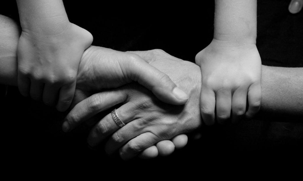 Three hands clasped: adult's hand in a handshake with another adult, a child's hand on top for support.