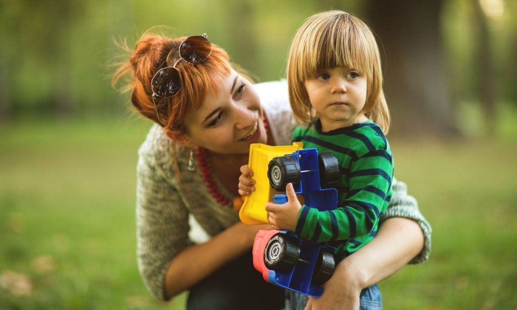 Woman crouching beside child, holding toy cars in a park.