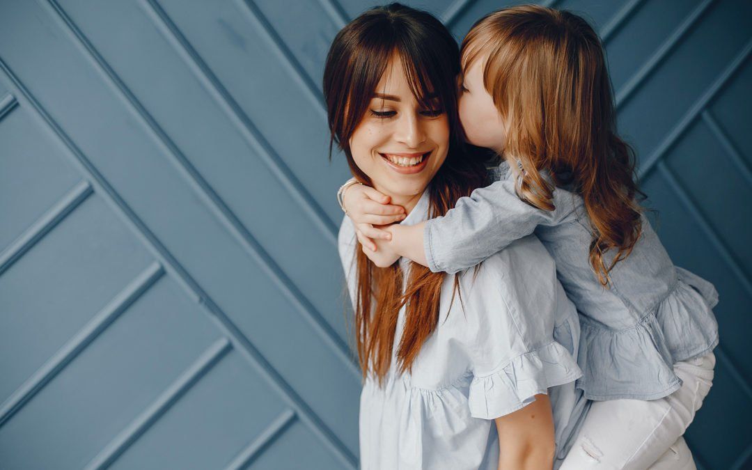 Woman smiling, child whispering in her ear, blue backdrop.