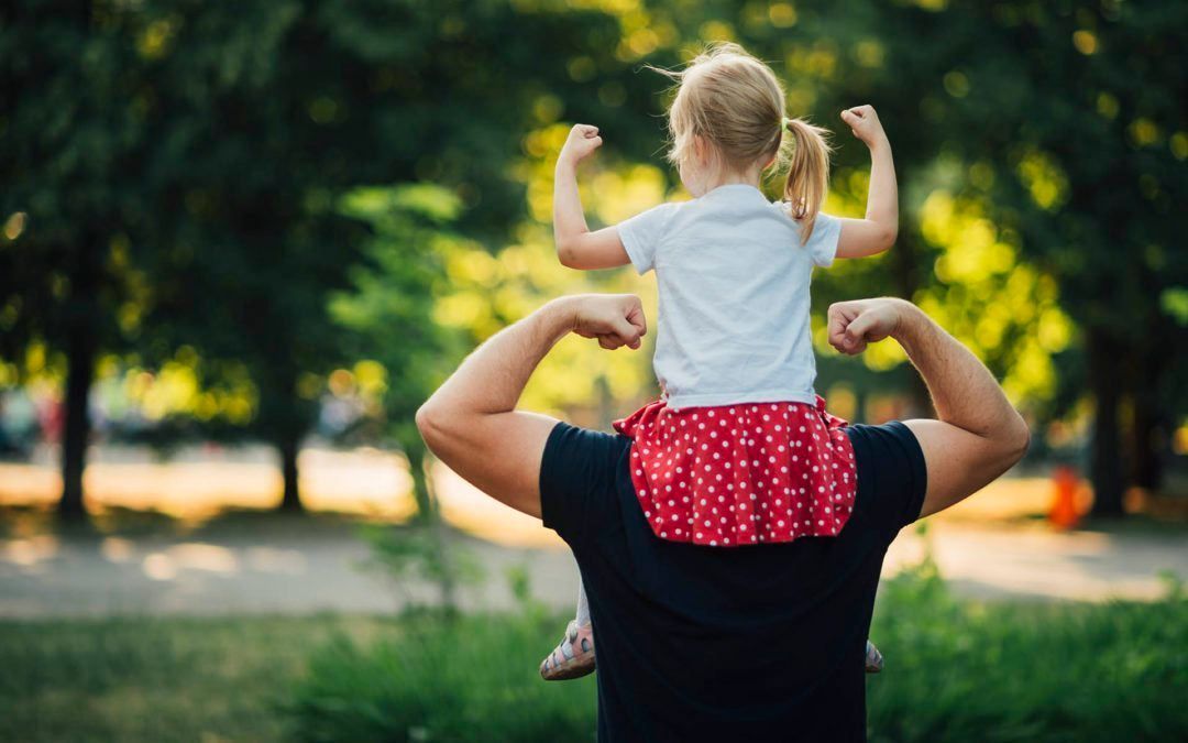 Child sits on person's shoulders, flexing arms. Outdoors, green foliage in background.
