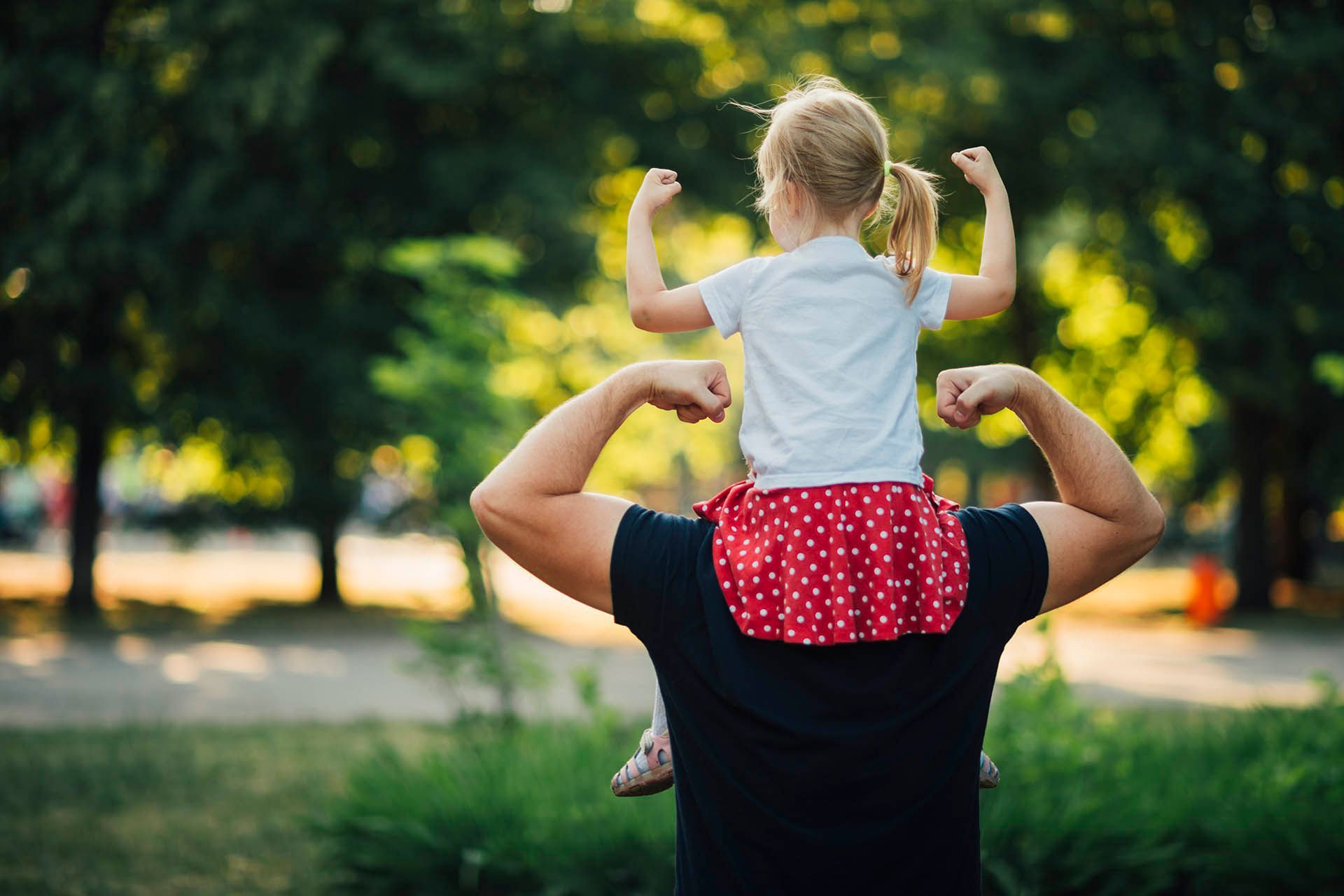 Child sitting on a person’s shoulders, flexing arms. Red and white skirt, green foliage in the background.