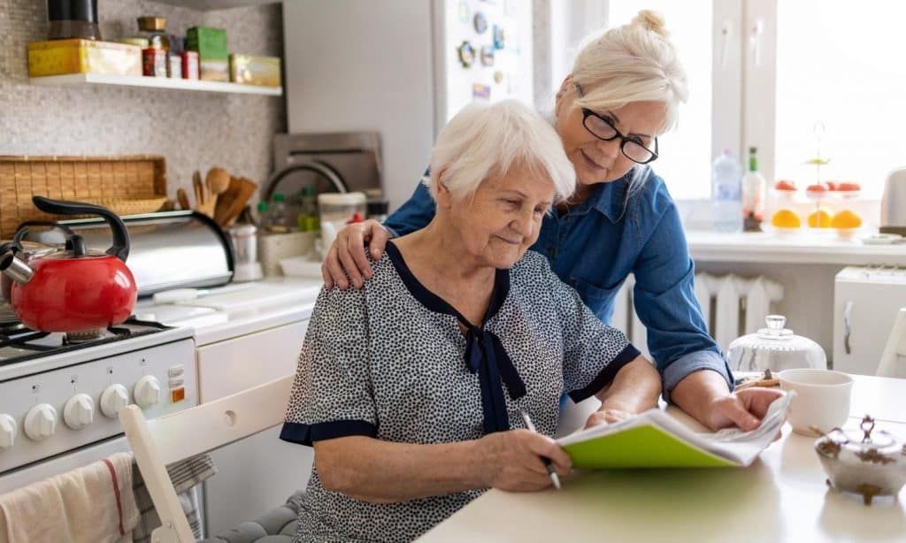 Two women in a kitchen, one assisting the other with a document.
