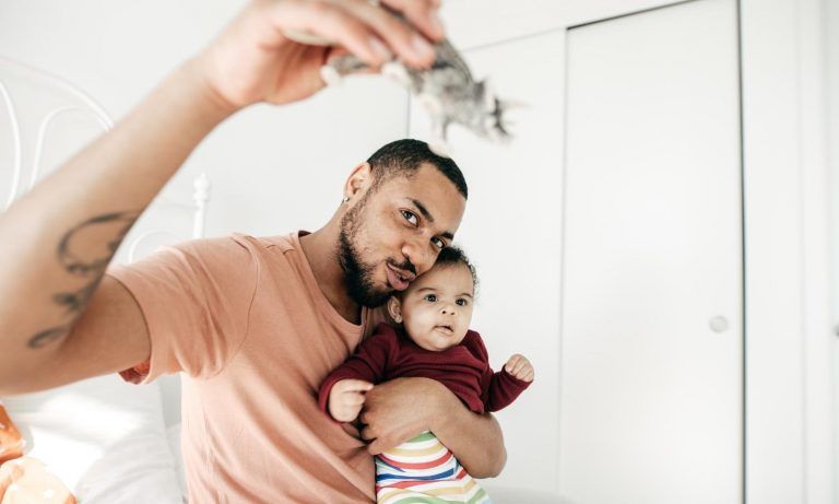 Man holding baby, showing toy. Both looking up; peach shirt, burgundy top, striped pants, white door.