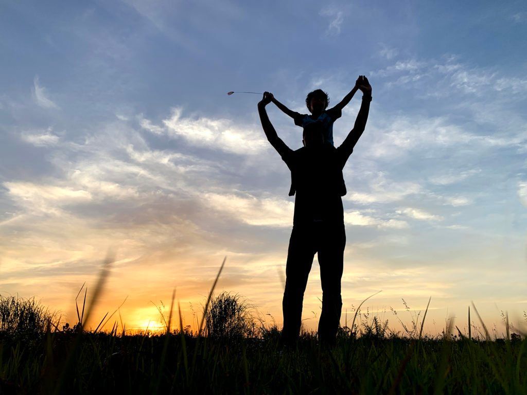 Silhouette of a person carrying a child on their shoulders, arms raised, against a sunset.