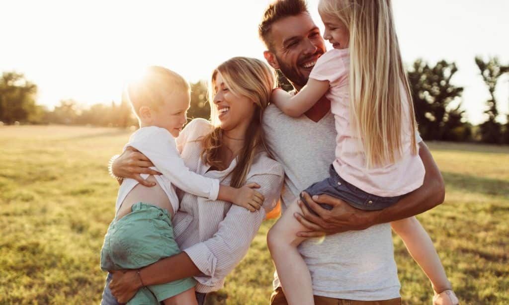 Family of four laughing, embracing in a grassy field; sunlight in background.