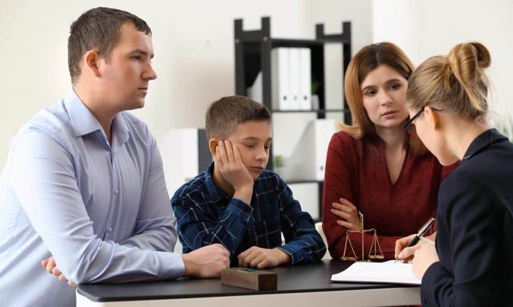 Family and lawyer at a table; child looks sad. Office setting; woman writes.