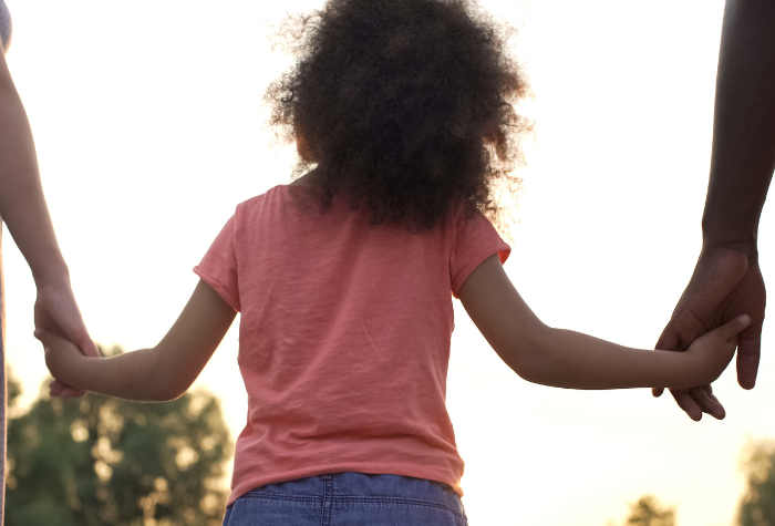 Child holding hands with two adults, silhouetted against a bright sky.