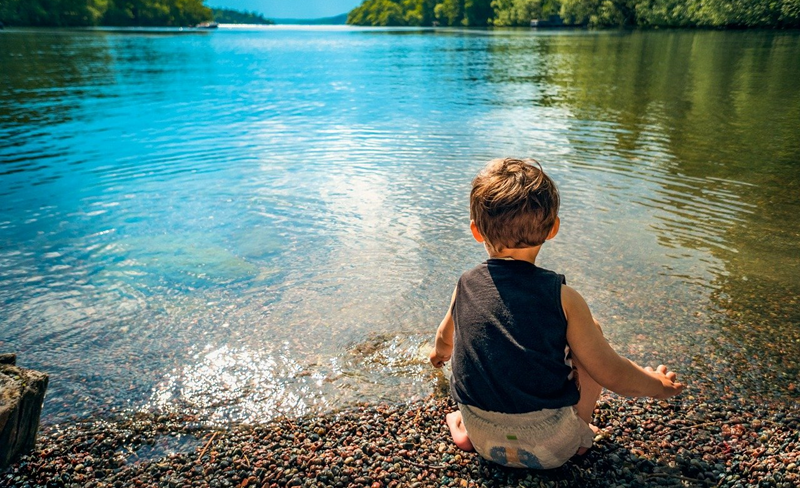 Child sitting at the edge of a lake, looking at the water. Blue water, green trees, sunny day.
