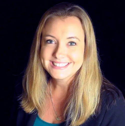 Woman with blonde hair smiling, wearing a teal top and black blazer against a dark background.