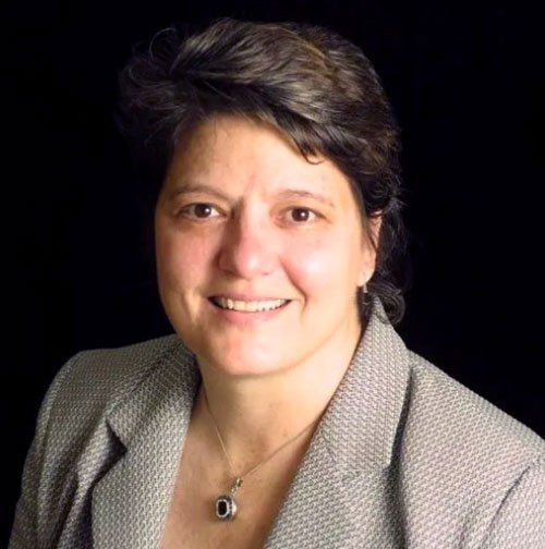 Woman with short dark hair smiling, wearing a patterned blazer and necklace, against a black background.