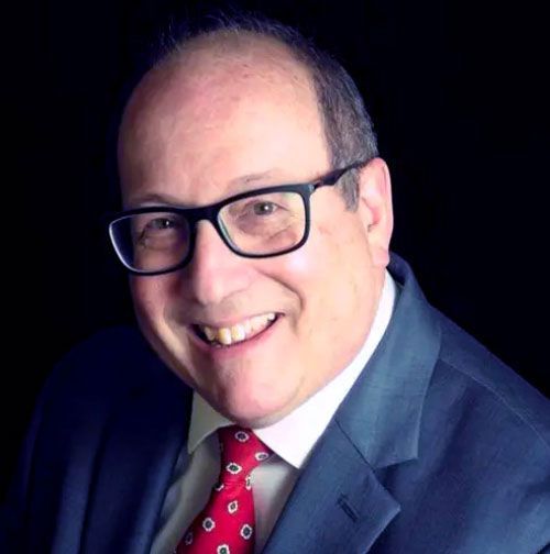 Man wearing glasses, suit, and red patterned tie smiles broadly against a black background.