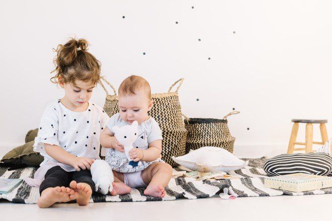 Two young children sit on a striped blanket with toys, next to woven baskets and a stool.