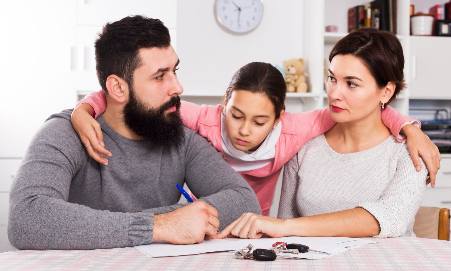 Family at table: Father signing document, daughter between parents with arms around them, mother looks on.
