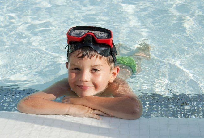 Boy in pool wearing goggles, smiling, resting arms on the edge of the pool.