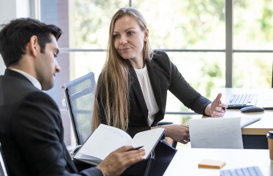 Man and woman in a meeting, reviewing documents at a table. Woman gestures, man writes.