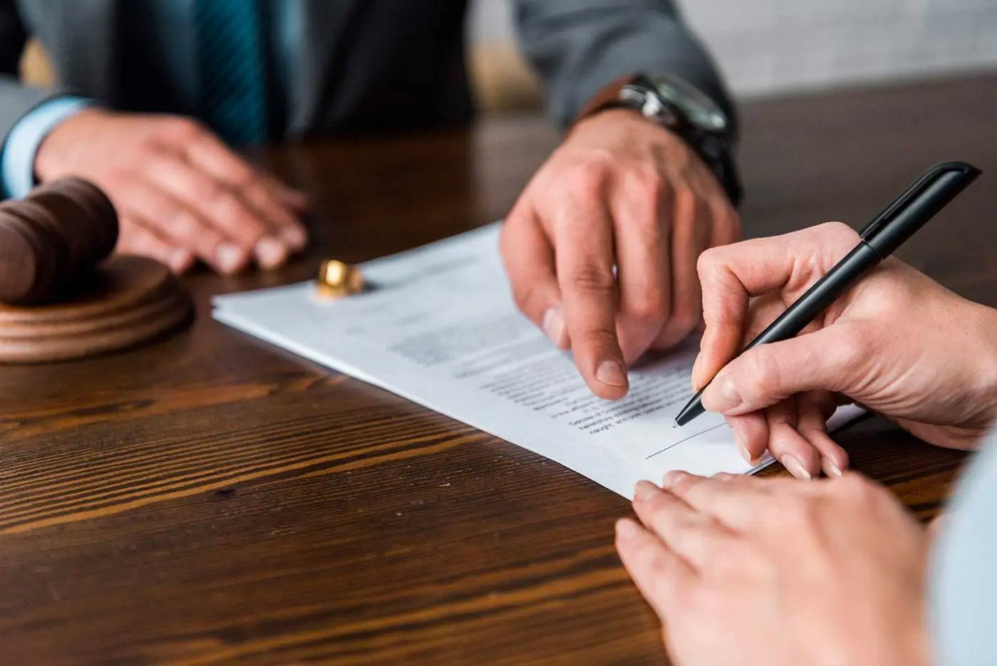 Hands signing a legal document on a wooden table, with a gavel and wedding ring visible.