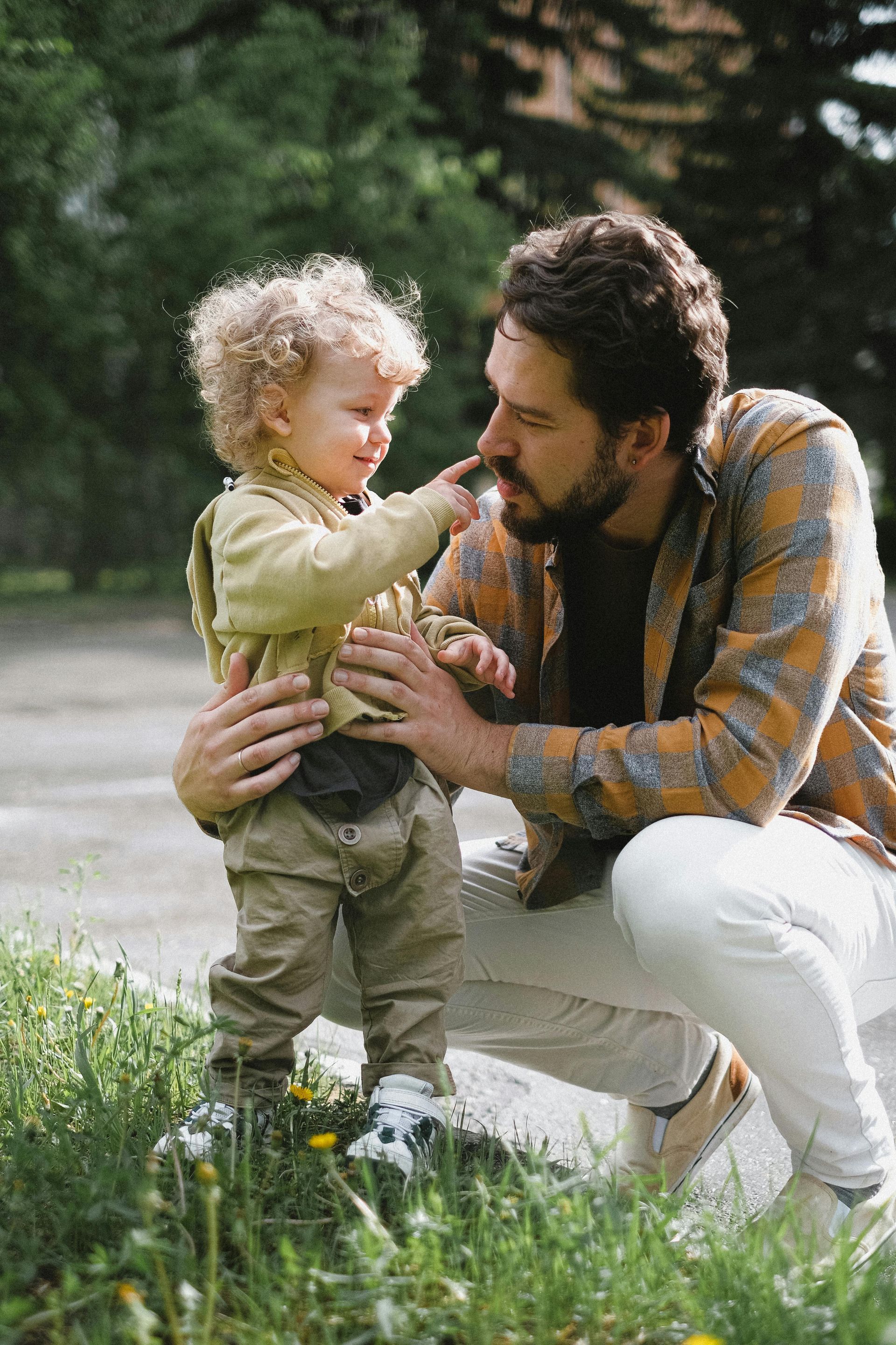 A child with curly hair touches a man's nose as he kneels in grass near a tree.
