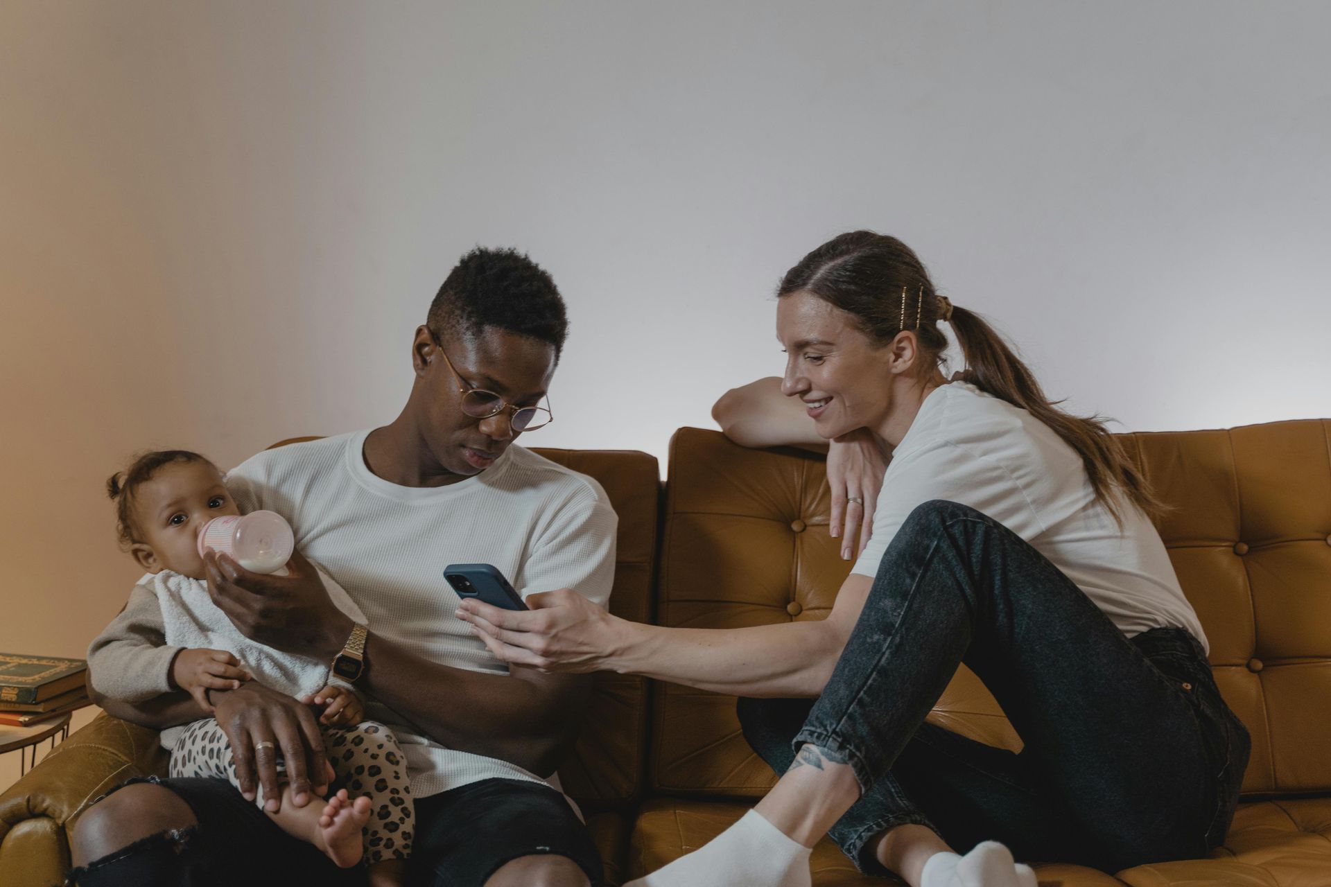 Family on a couch: one parent feeding a baby while the other looks at a phone and leans in.
