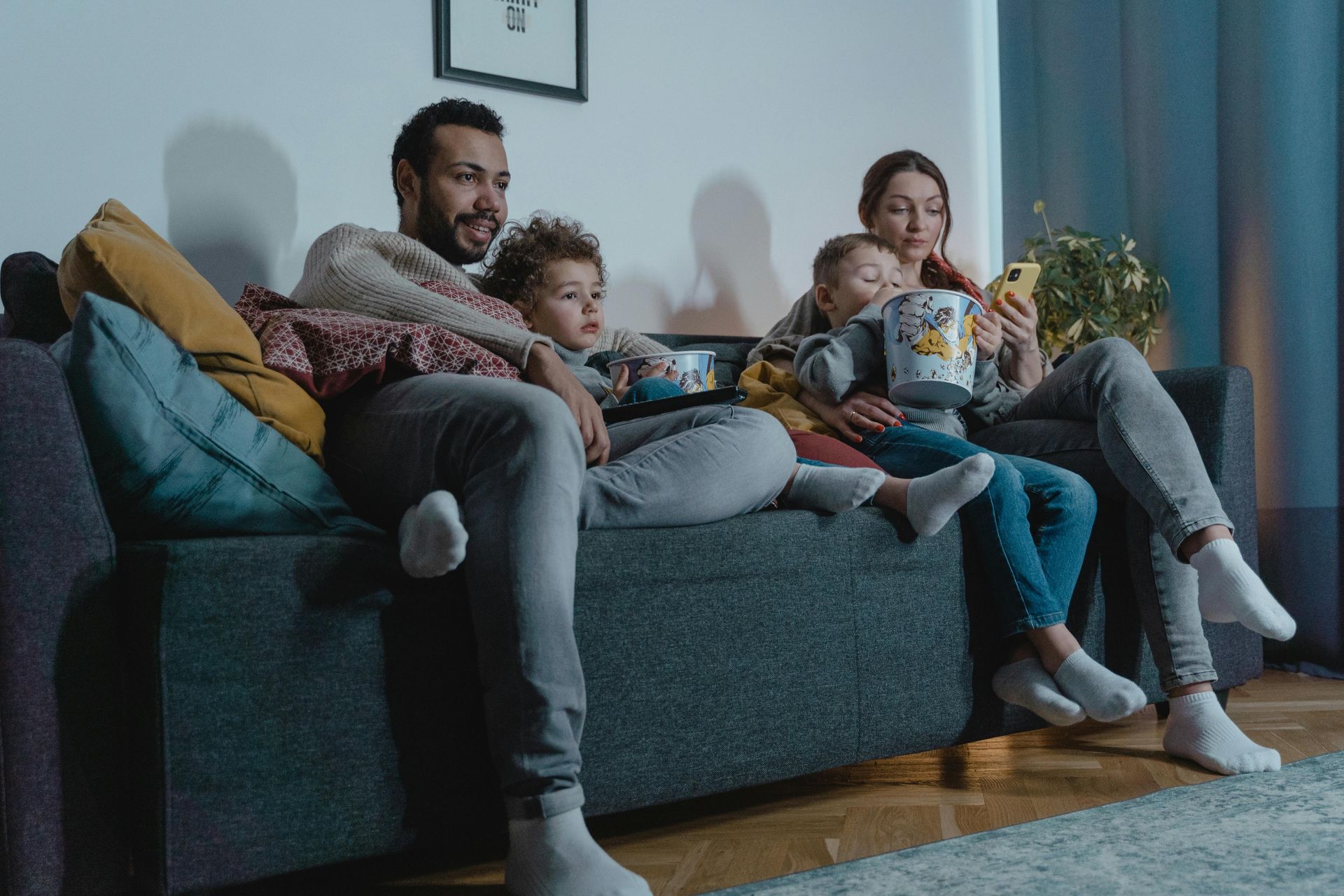 Family on a couch watching TV, eating popcorn. Dark room, neutral colors, cozy atmosphere.