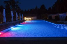 Illuminated blue swimming pool at night with lounge chairs and closed umbrellas nearby