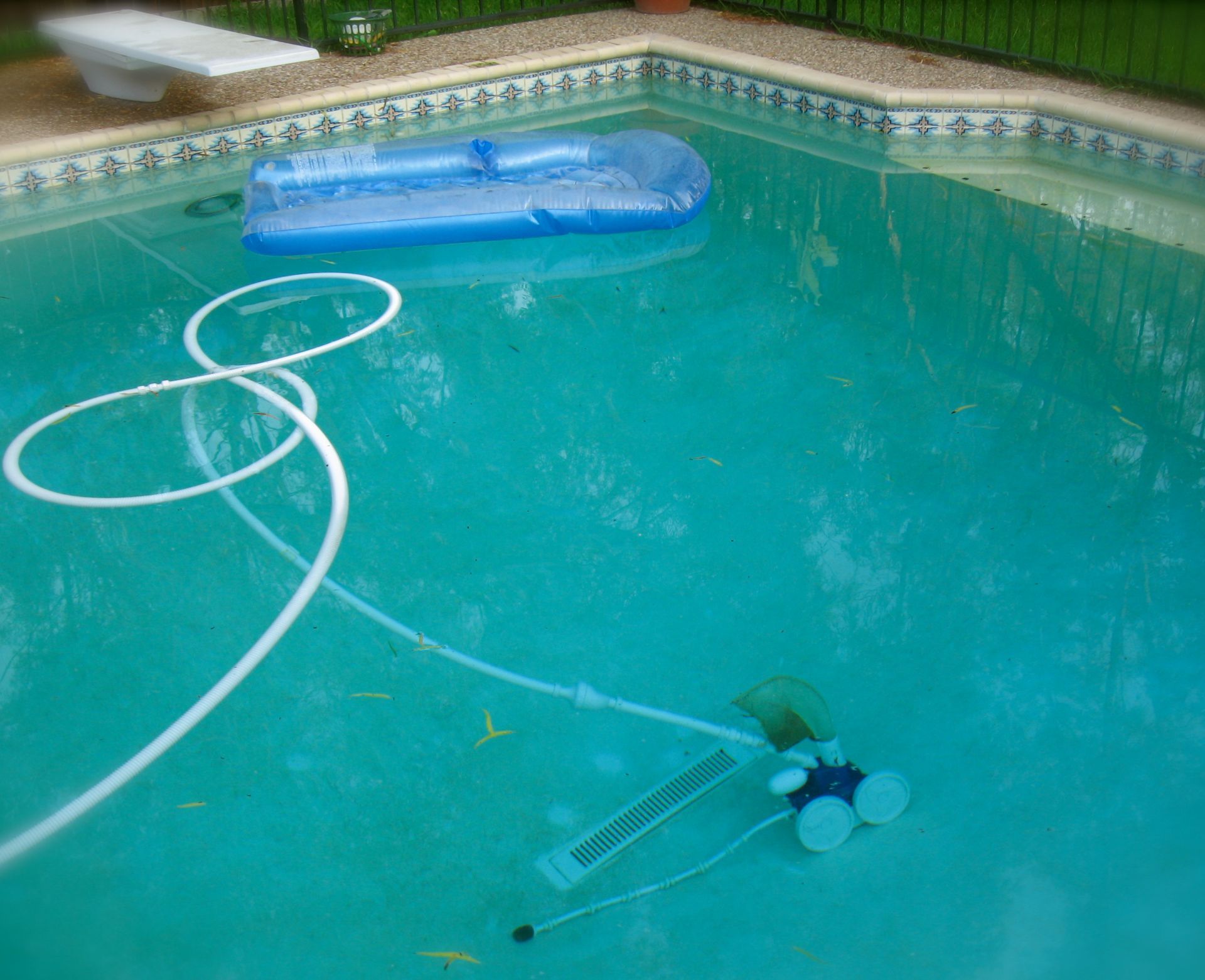 Backyard swimming pool with a floating blue cover, pool hose, and cleaning equipment in the water