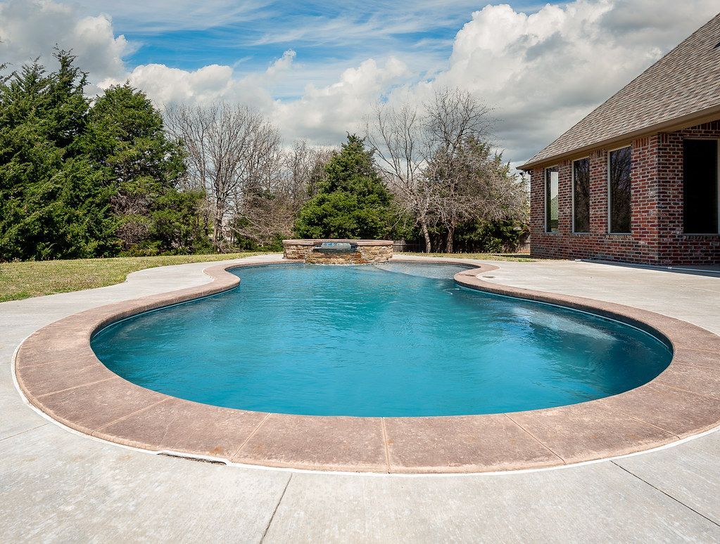Curved backyard swimming pool with blue water beside a brick house and trees