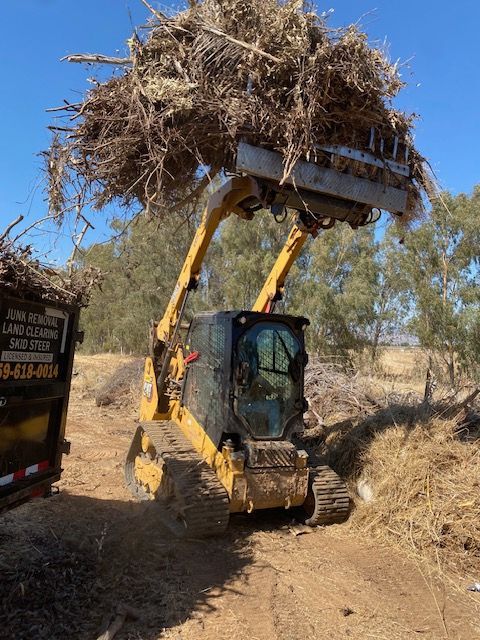 Yellow excavator lifting a large bundle of dry brush on a dirt path in a rural field
