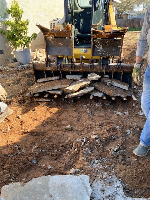 Excavator clearing dirt in a backyard, with broken stone slabs and a person standing nearby.