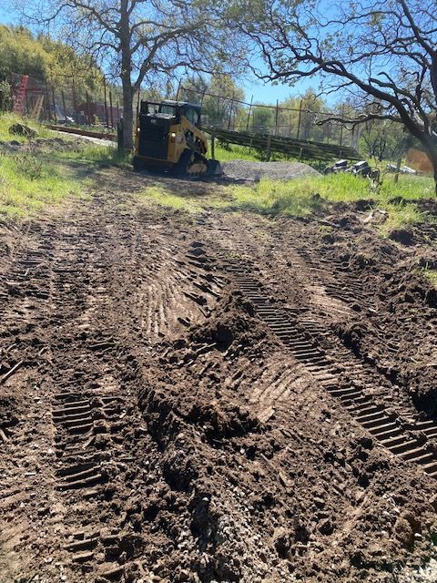 Mud-covered dirt road with deep tire tracks leading to a bulldozer in a grassy, tree-lined field