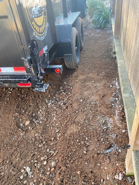 Utility trailer on a narrow dirt path beside a wall and fence, with rough uneven ground.