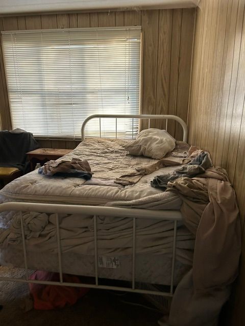 Unmade bed with clothes and pillows in a wood-paneled bedroom beside a window with blinds