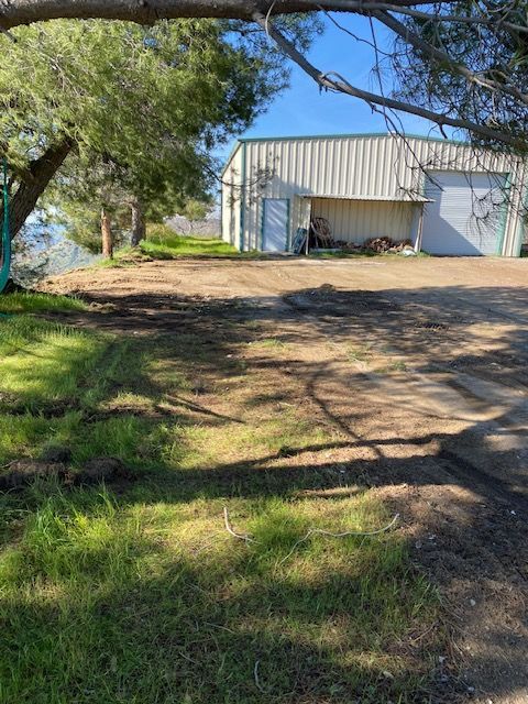 Shaded dirt yard with trees and grass in front of a white metal shed under a blue sky