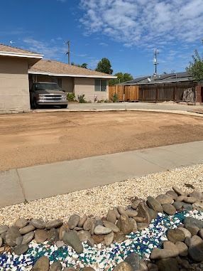 Single-story house with a carport and dirt driveway, bordered by rocks and blue-white landscaping stones.