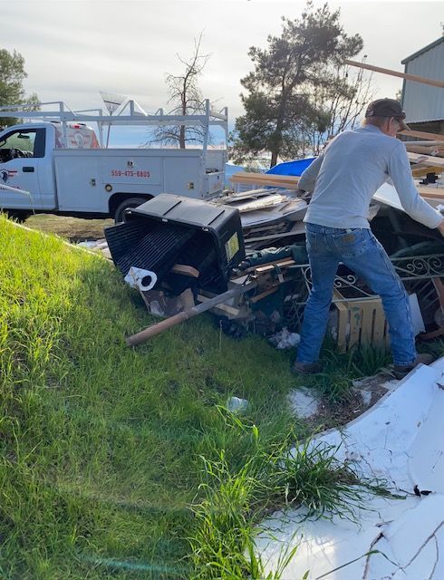 Man standing in a messy yard beside a tipped black trailer, with a white utility truck in the background.