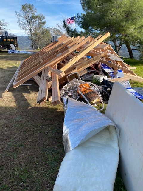 Collapsed wooden roof structure on a lawn beside a white tarp, with debris and an American flag in the background