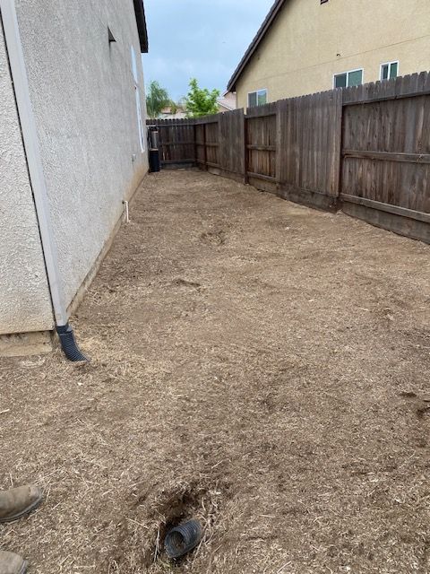 Narrow dirt side yard between a white house and wooden fence, with a small drain near the foreground.
