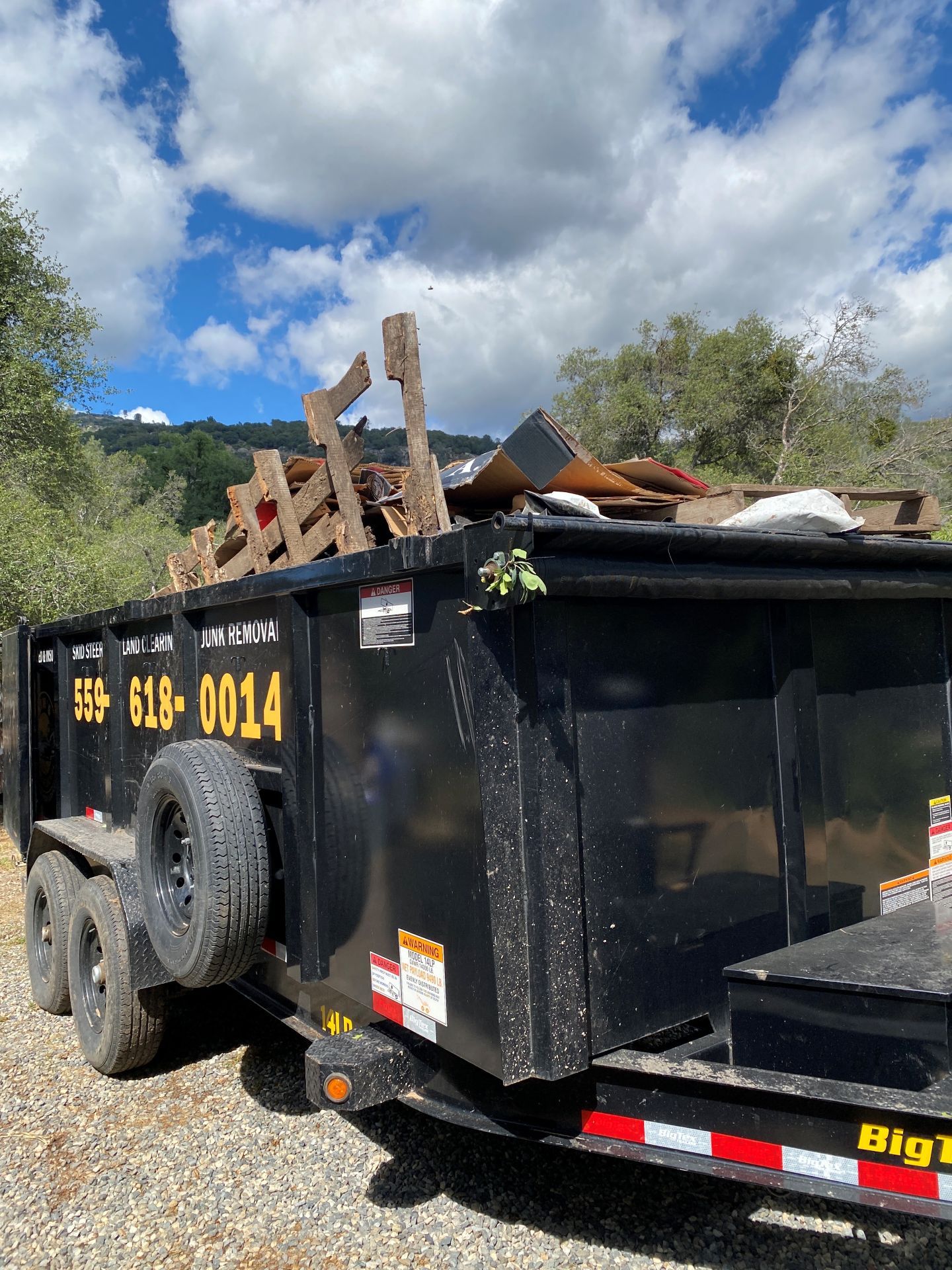 Black dump trailer filled with wood debris, parked outdoors under a cloudy sky.