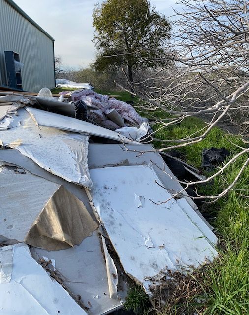 Pile of broken gray roofing or siding panels beside a building and tree in a grassy yard