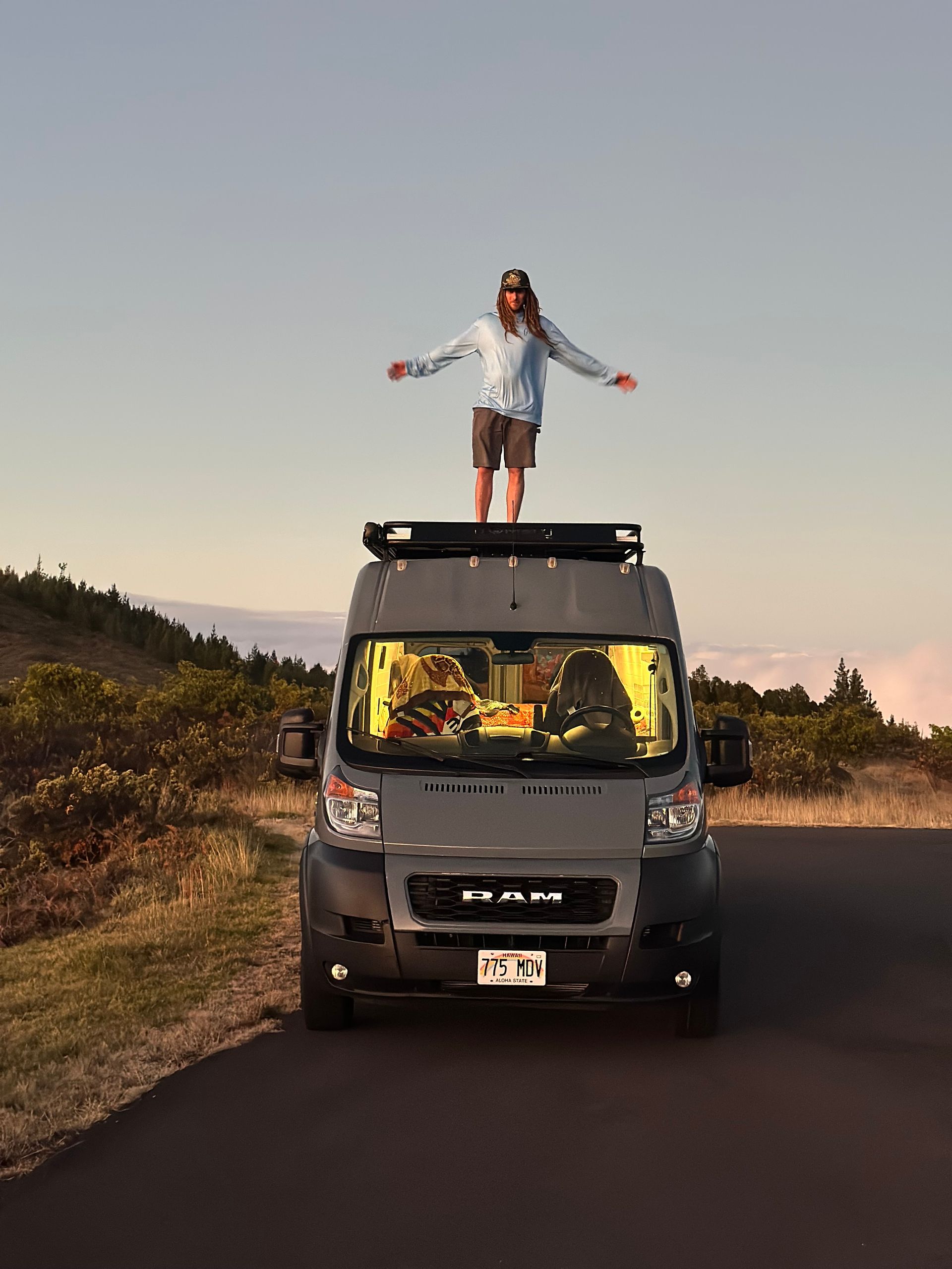 Traveler standing on camper van roof at scenic roadside viewpoint in Maui during sunrise or sunset