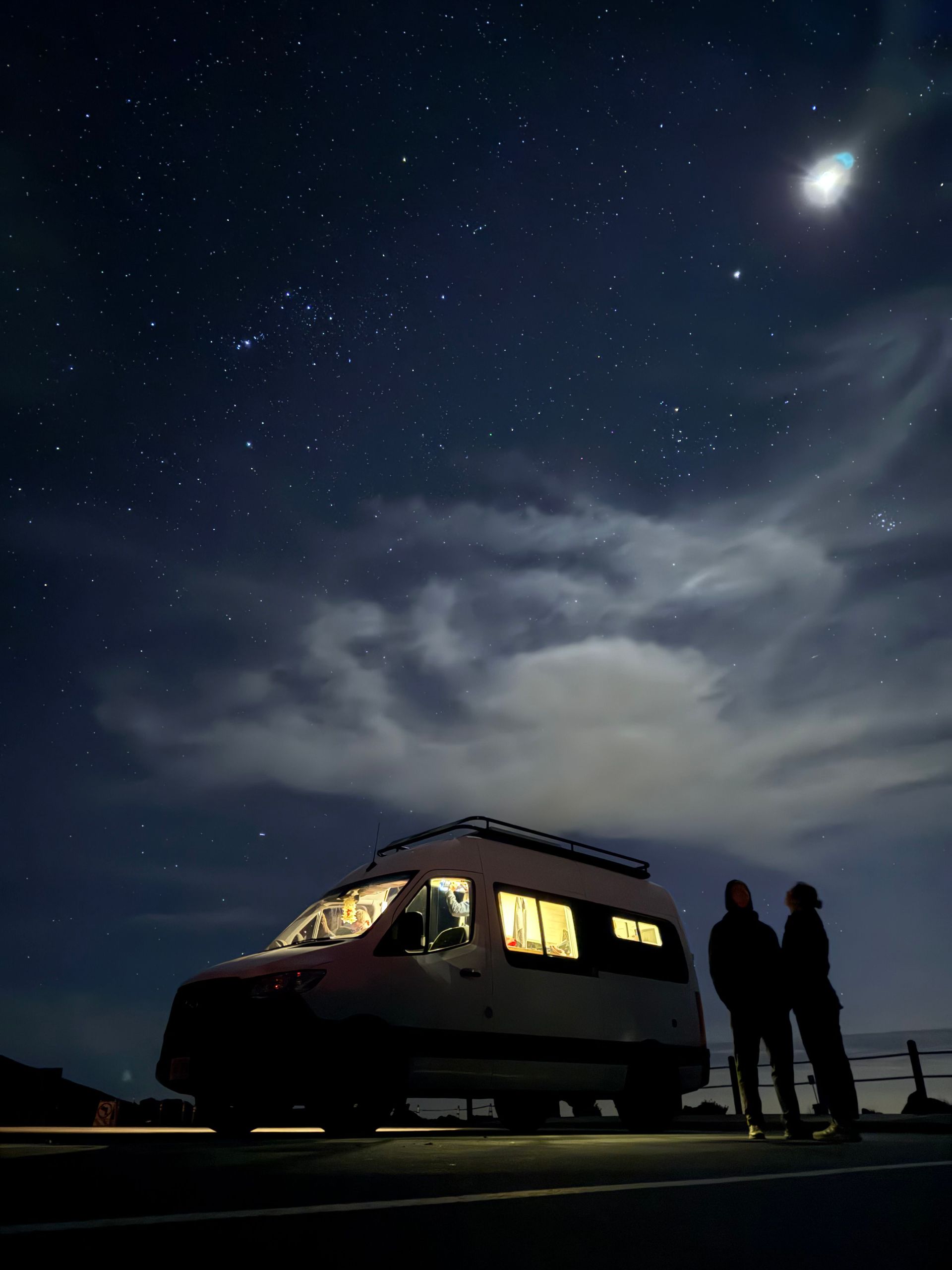 Van under starry night sky; two figures silhouetted nearby. Interior lights glow.