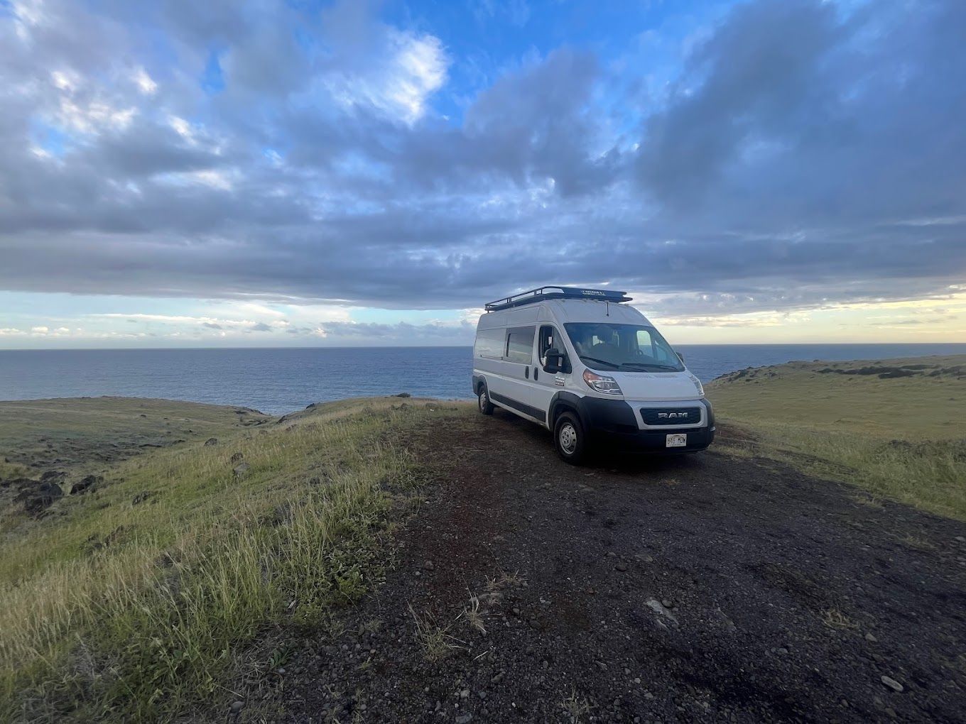 A white camper van parked on a grassy cliff overlooking the ocean under a dramatic cloudy sky at dusk.
