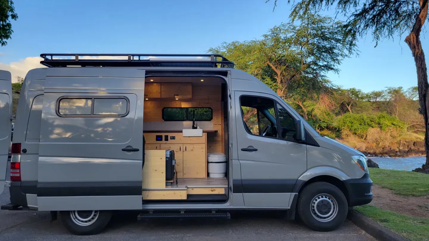 Gray camper van parked by a grassy riverbank, side door open to a compact wooden interior.