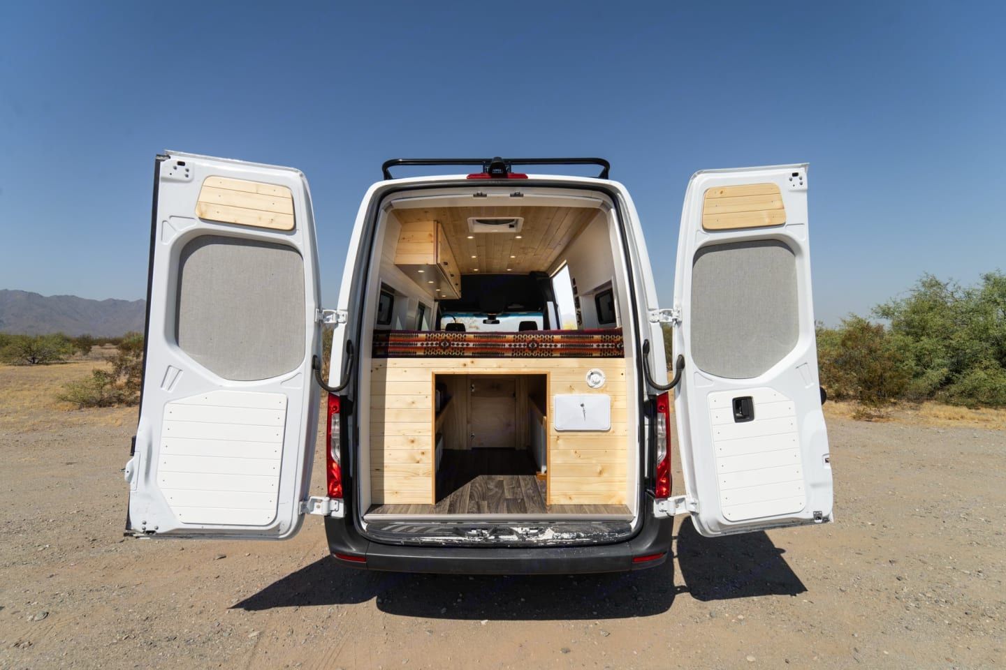 Open rear of white van revealing a simple wooden camper interior in a desert setting