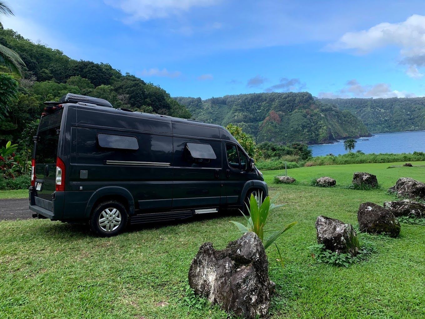 Gray camper van with side door open, parked in a wooded area with a person sitting at the entrance.