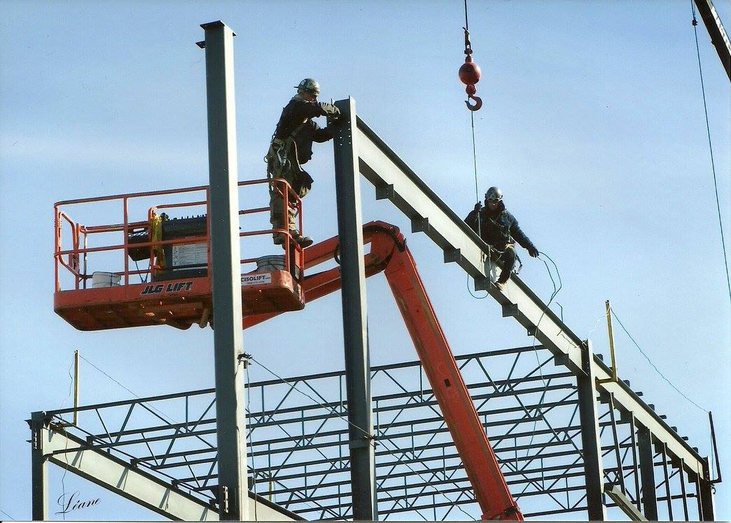 Deux ouvriers, juchés sur des plateformes élévatrices, assemblent une structure en acier sur un chantier de construction.
