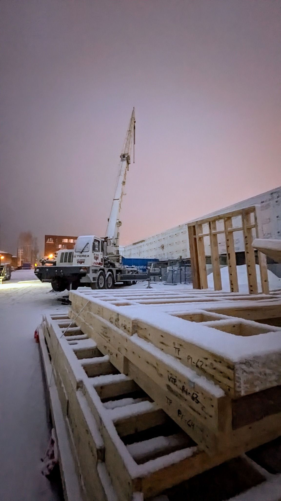 Chantier enneigé avec une grue soulevant des matériaux à côté de poutres en bois empilées au crépuscule